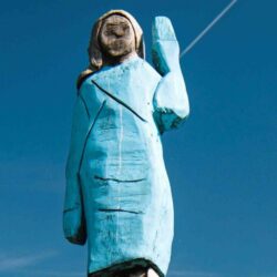 wooden sculpture of a badly rendered woman wearing a blue skirt suit with left arm raised in greeting
