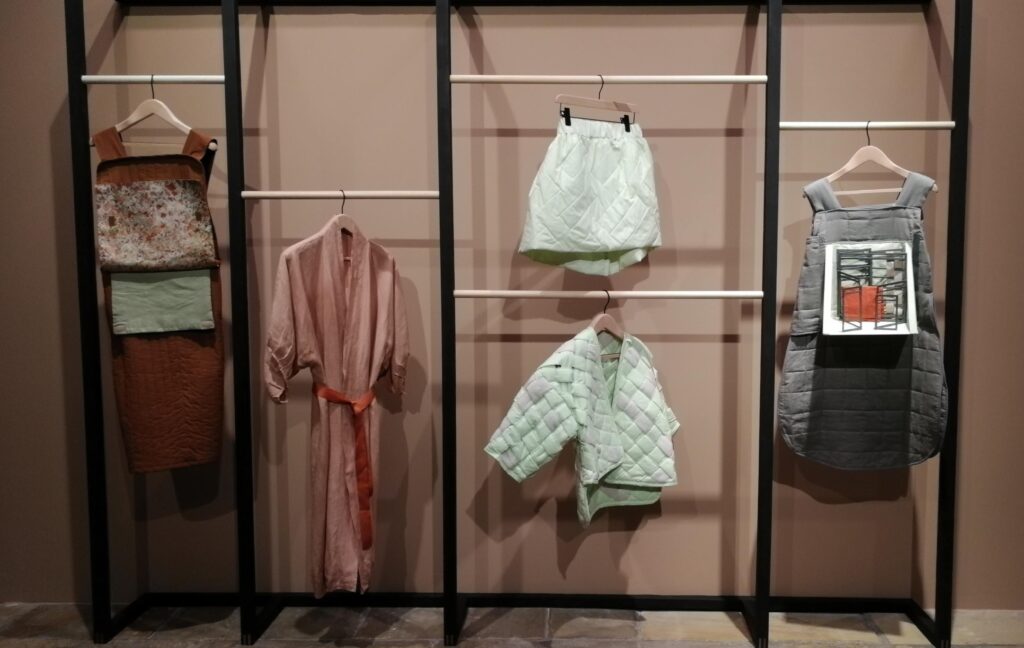 Rails of clothing from Emily Speed's "Flatland". From left-right: a brown tabard with flat layers, a pink kimono, a green thick-weave skirt and jacket, and a grey tabard with a model set built onto the chest.